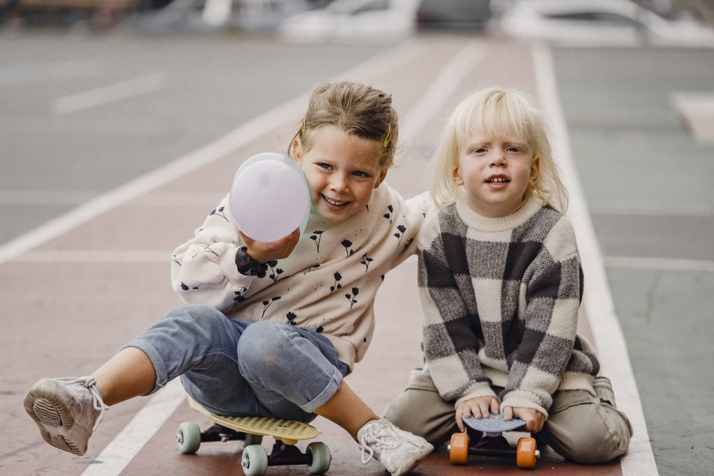 Full body of smiling children in casual wear sitting on skateboards on asphalt road in daytime